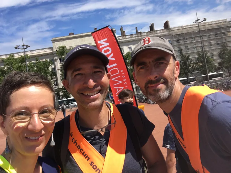 Selfie de Camille, Marc et Thomas. Les trois sourient. Ils sont habillés de gilet fluo. Il fait grand soleil. En arrière plan, on devine la Place Bellecour à Lyon.