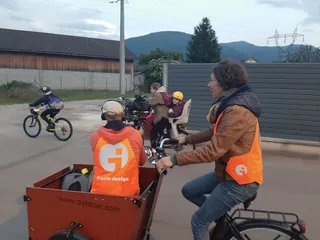 Camille conduit un vélo cargo tandis que Marc, dans la caisse avant filme. En arrière plan, une maman et ses enfants font du vélo.
