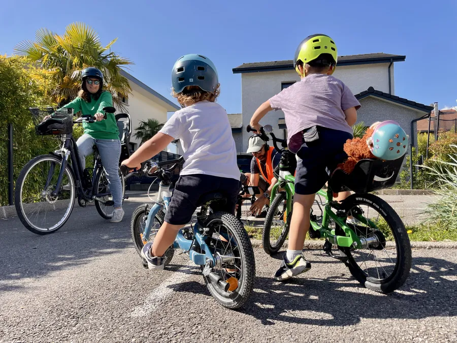 Sous un soleil de printemps, deux petit garçons enfourchent leurs petits vélos bleu et vert. Ils sont de dos et regardent une adulte, à vélo, qui les accompagne.